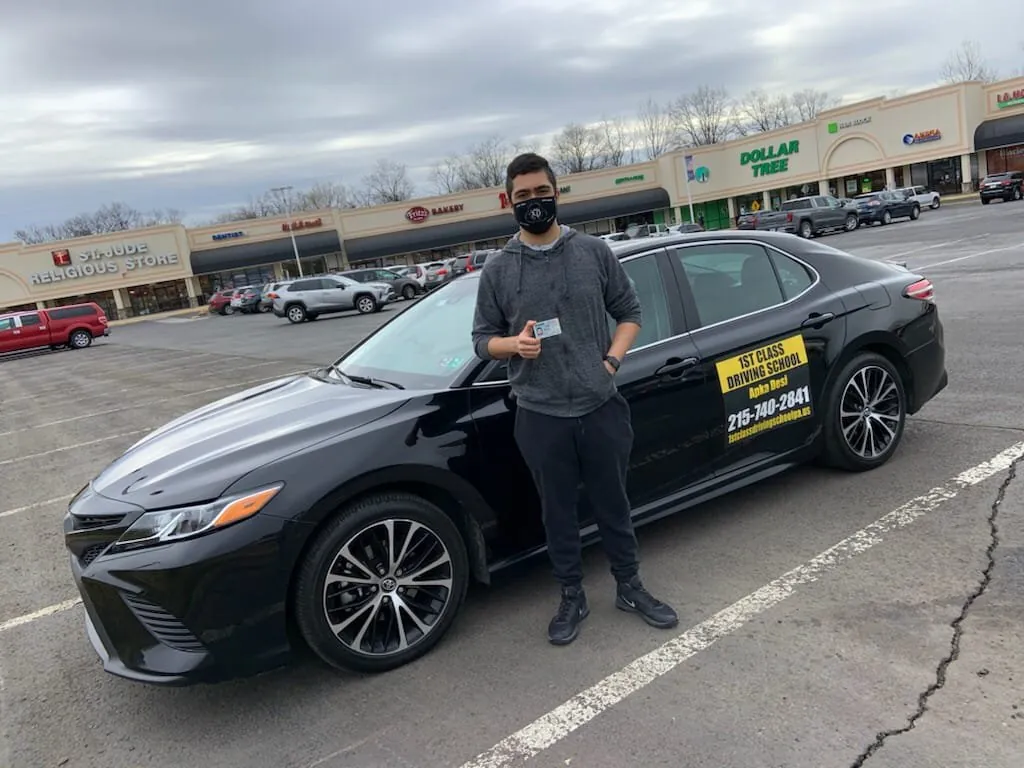 Student standing beside the black dual-control training car in a shopping-center parking lot